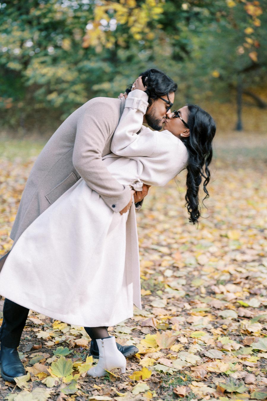 A couple sharing a romantic kiss in a park during autumn, surrounded by fallen leaves and lush greenery in the background.
