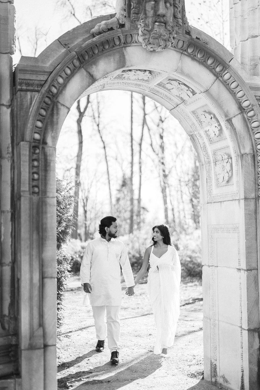 A couple holding hands while walking under a vintage stone archway in a forest setting, dressed in traditional attire.