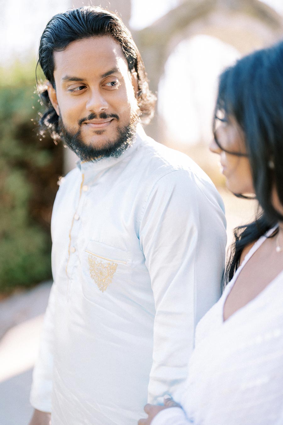 A man in a traditional white kurta with embroidered detailing is looking affectionately at a woman in a white outfit, outdoors on a sunny day.