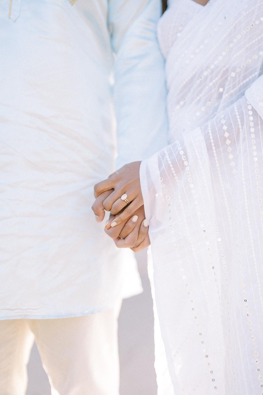 Close-up of a couple holding hands, showcasing wedding attire with intricate embroidery. The woman's hand features a prominent engagement ring, emphasizing love and commitment.