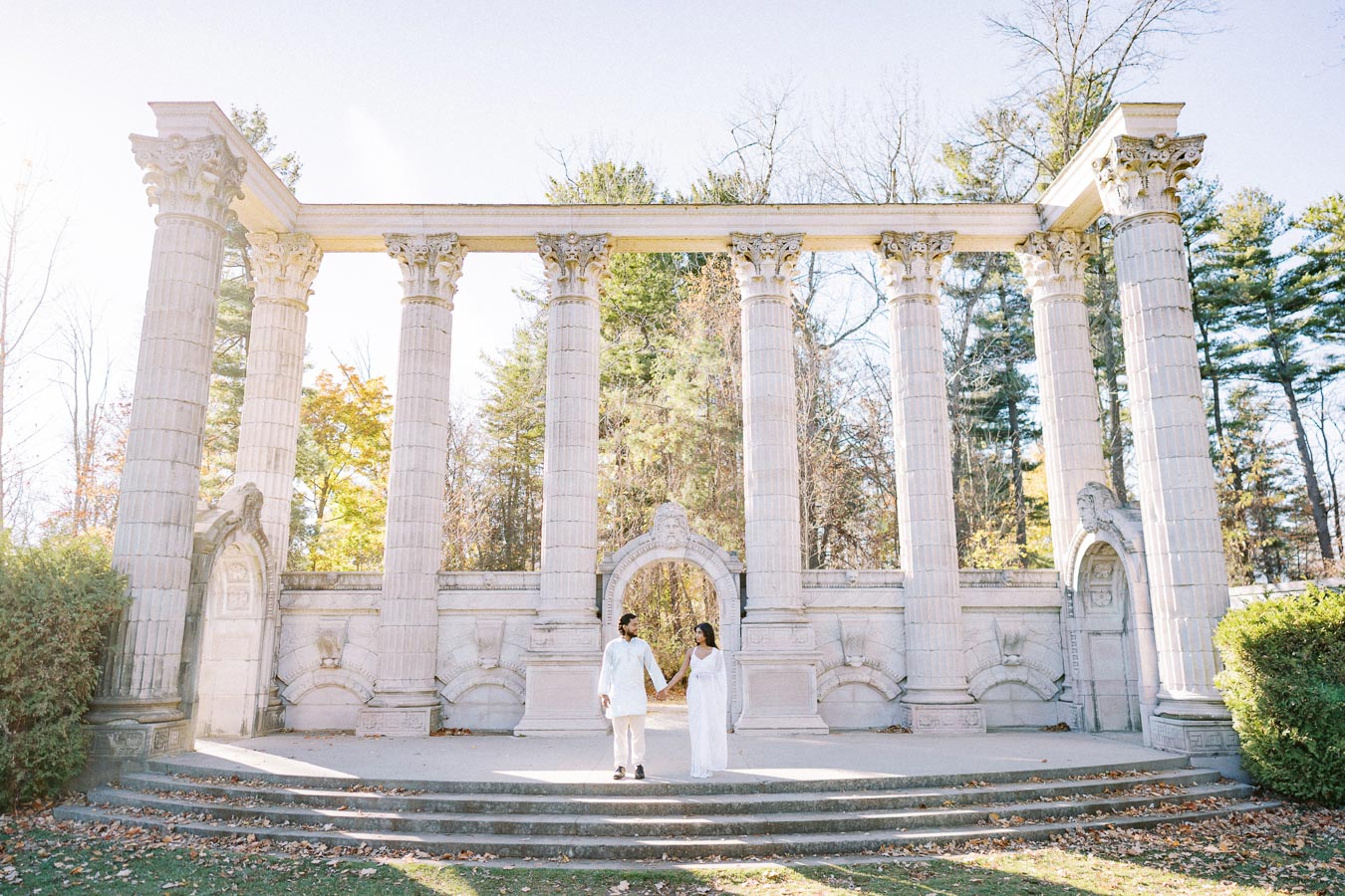 A couple holding hands in front of grand architectural columns and arches at an outdoor location, surrounded by lush greenery, under a clear blue sky.