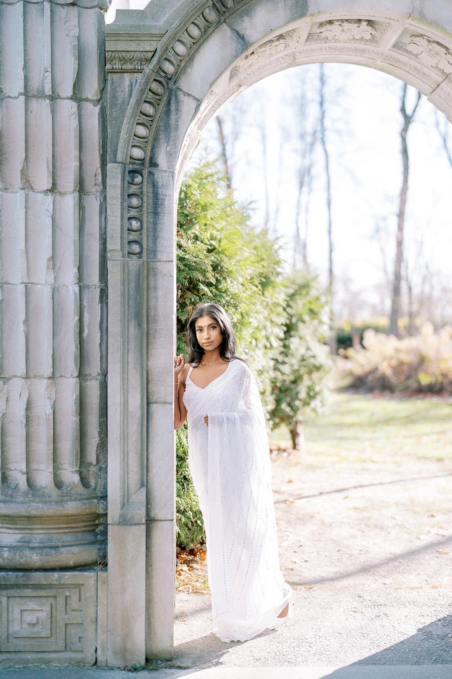 A woman in a white sari stands gracefully by an ornate stone archway in a sunlit garden.