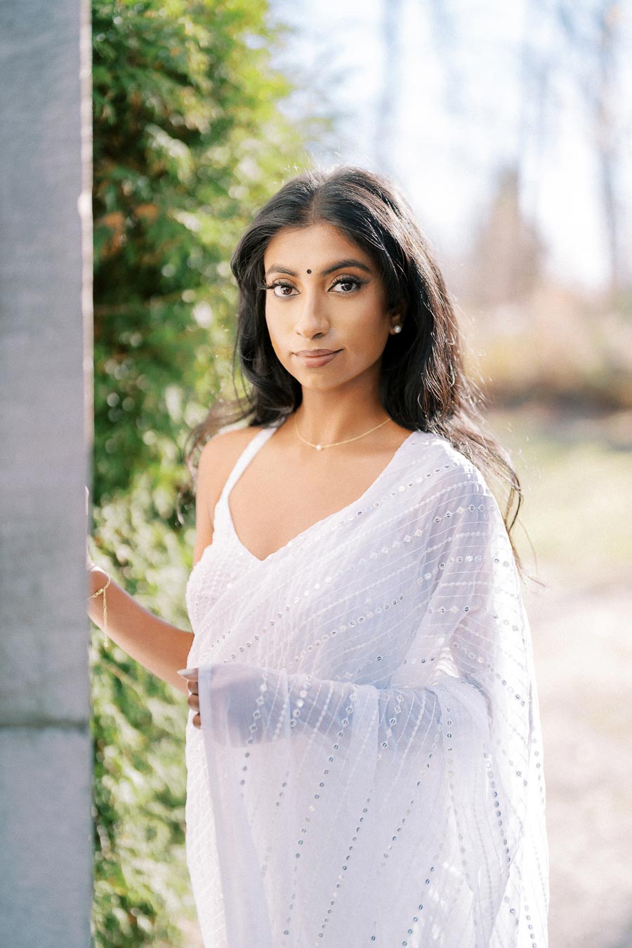 Woman in a white sari standing outdoors with a green background, showcasing traditional elegance and cultural attire.