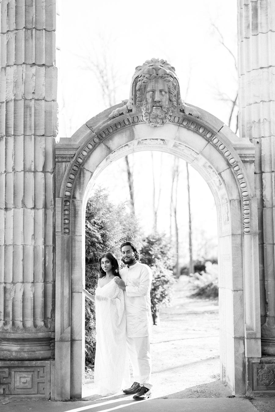 A couple posing elegantly under a stone archway adorned with classical sculpture, surrounded by greenery, in a serene outdoor setting.