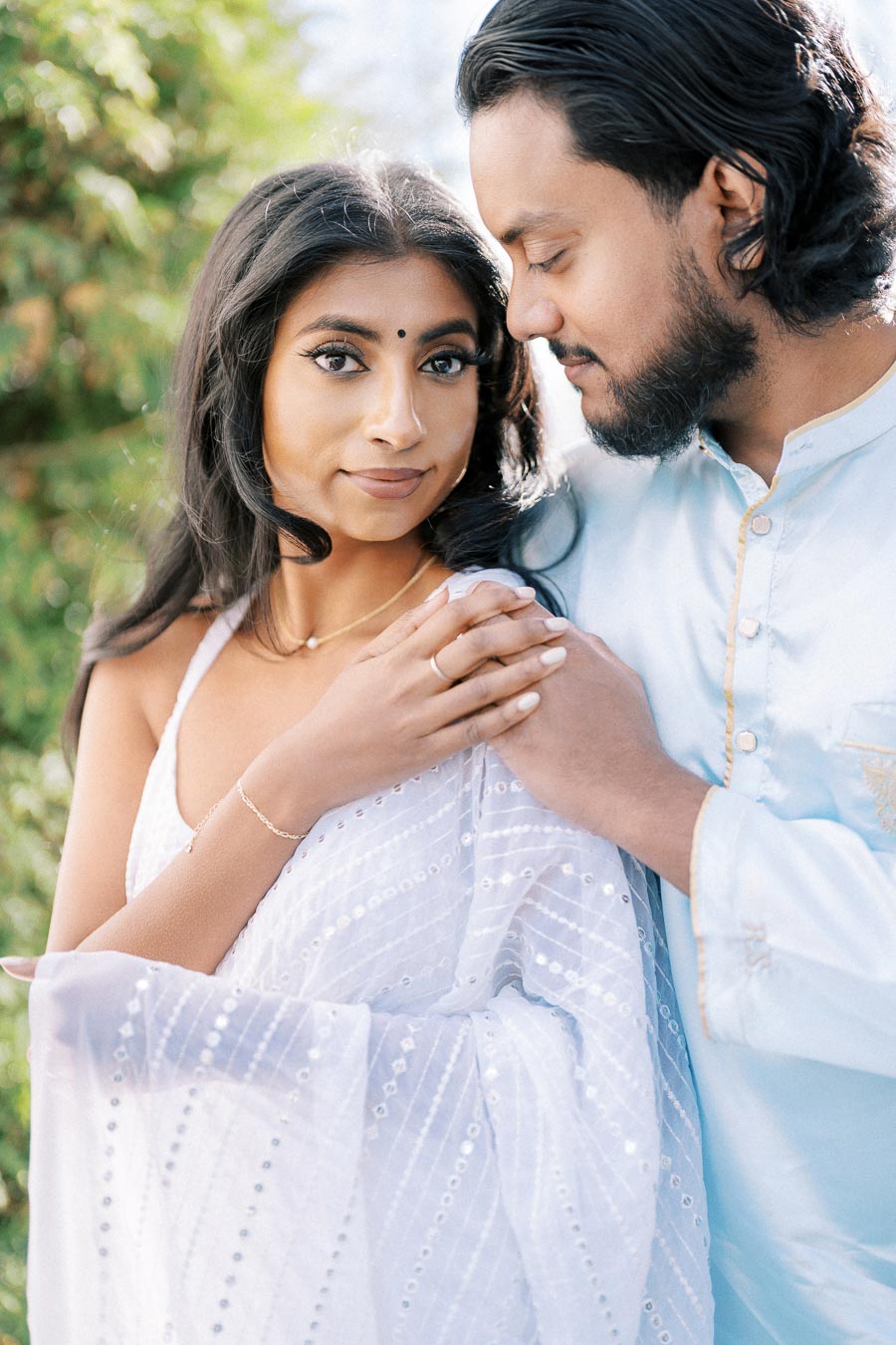 A couple wearing elegant traditional attire standing outdoors, with the woman in a white sari and the man in a light blue kurta, sharing a tender moment amidst a green, natural background.