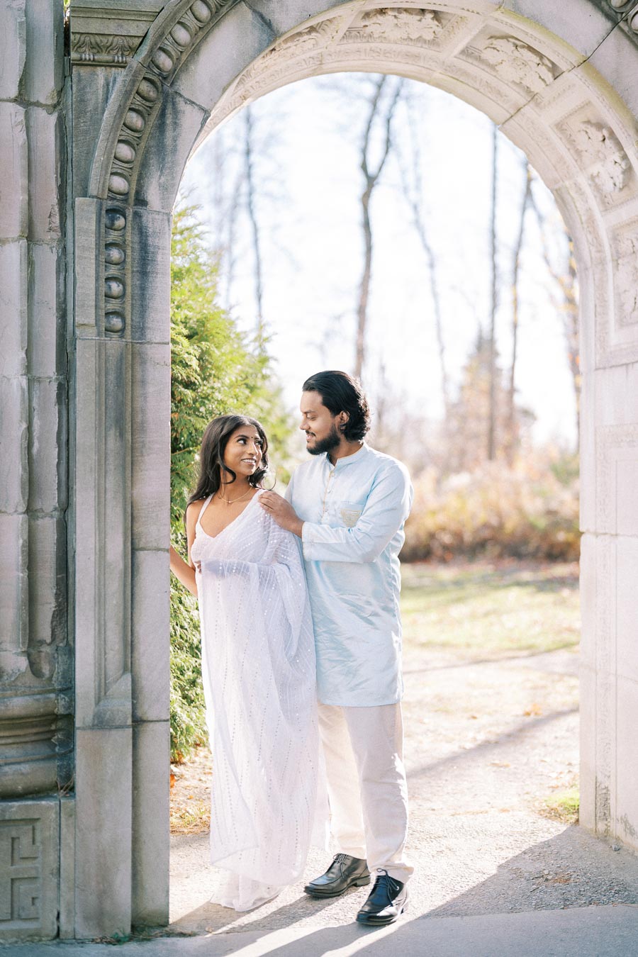 A couple in traditional attire share a tender moment under a historic stone archway, surrounded by lush greenery and a sunlit path.