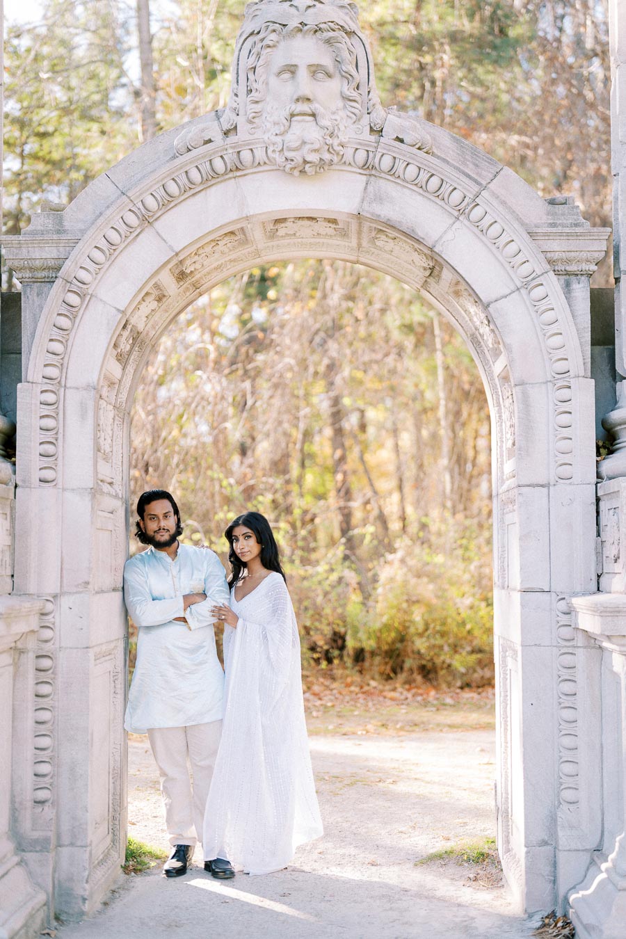 Elegant couple in traditional attire posing under a decorative stone archway in a serene park setting.