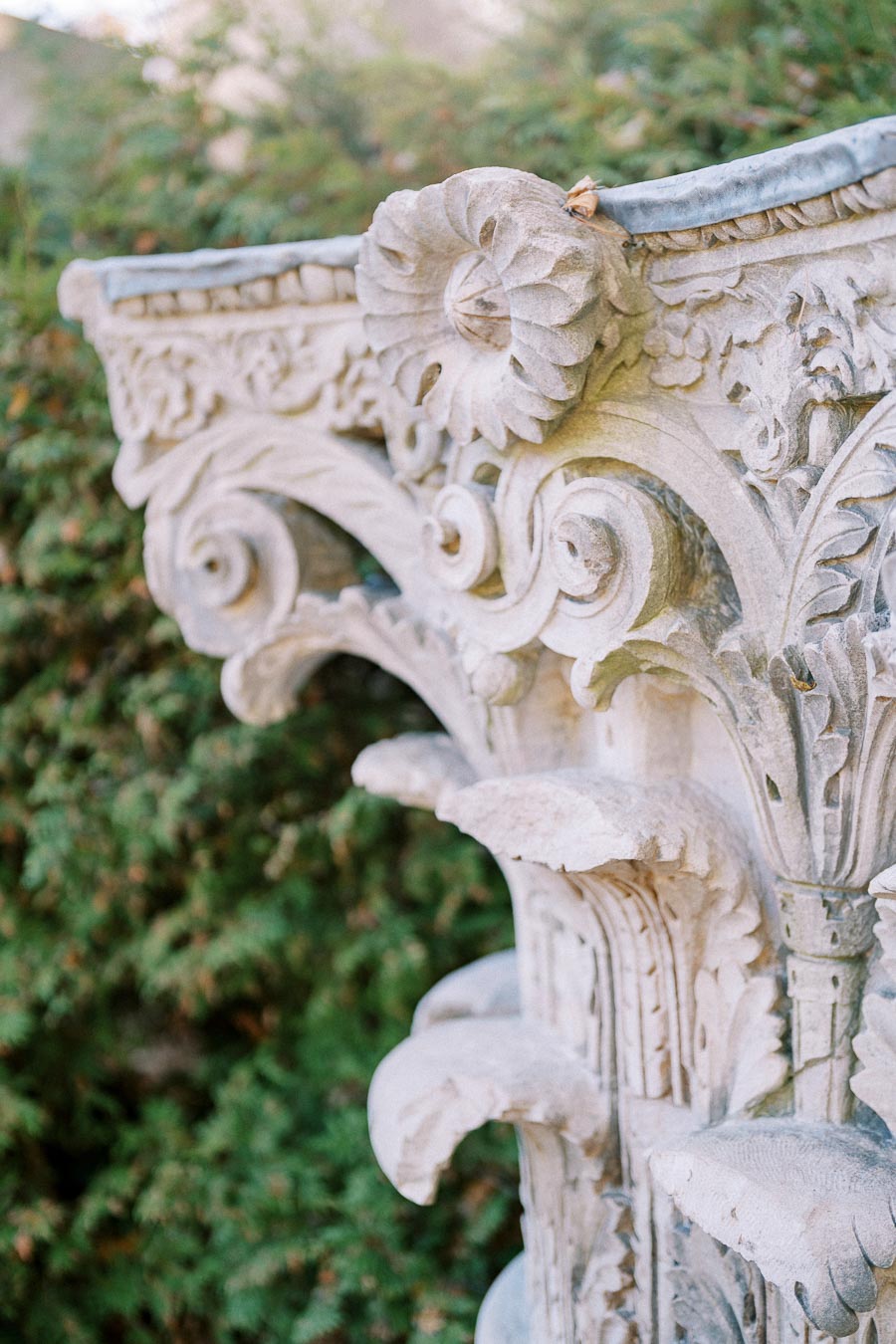 Ornate stone column capital with intricate carvings and decorative floral patterns, set against a lush green background.