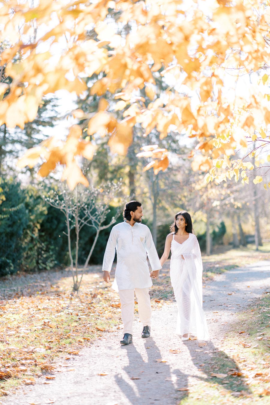 A couple in traditional white attire walking hand in hand along a scenic path covered in fallen autumn leaves, with golden foliage overhead in a serene park setting.