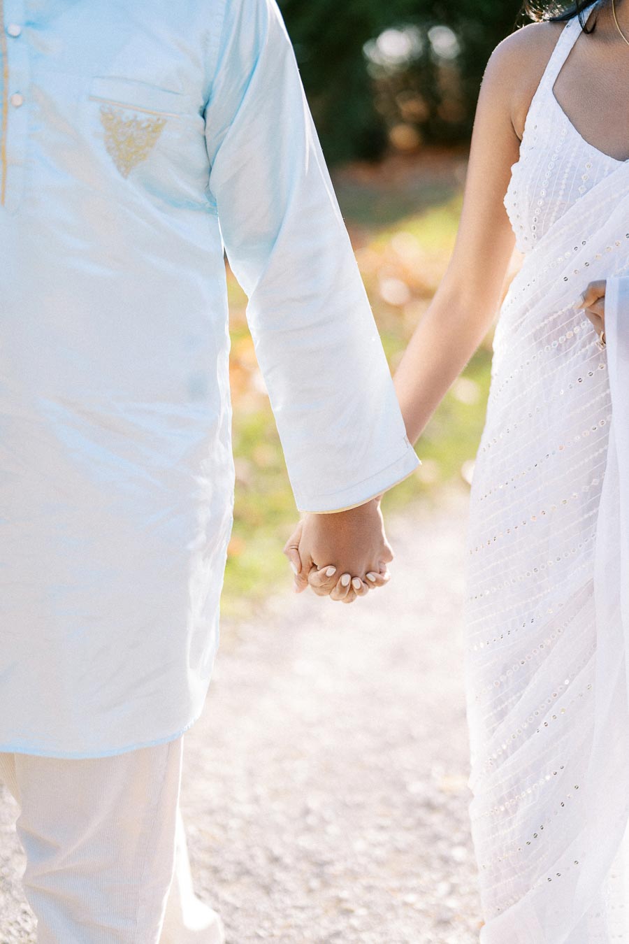 A couple holding hands, dressed in elegant traditional attire, walking on a sunlit path.