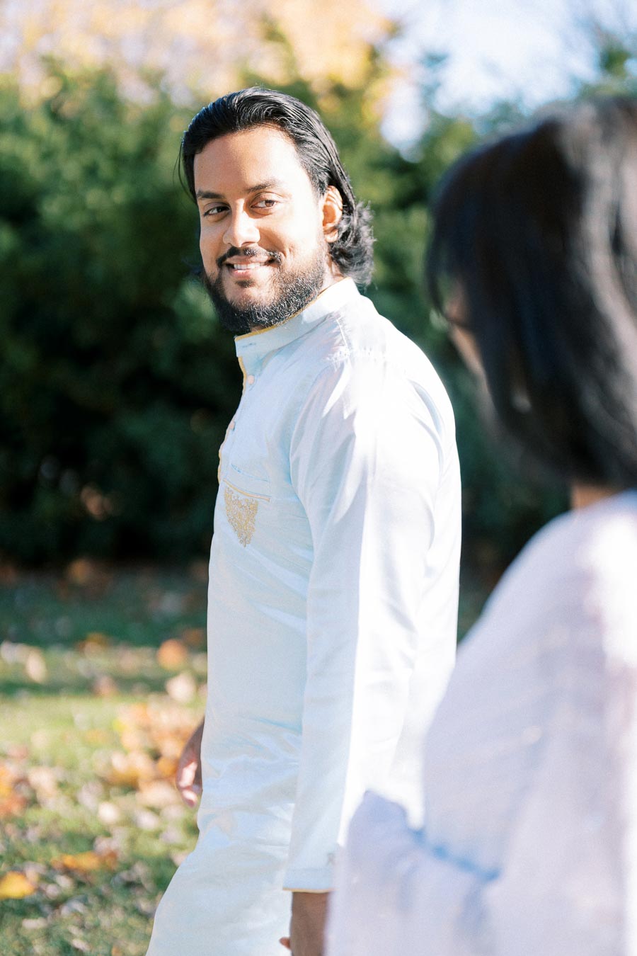 A man in a white traditional outfit walking outdoors in a sunlit garden setting, with trees and fallen leaves in the background.