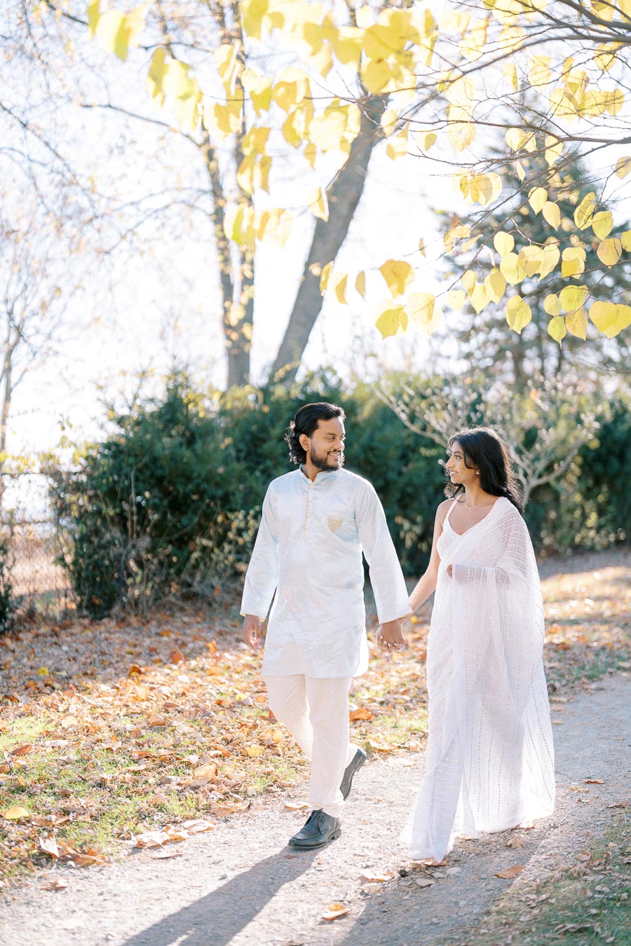 A couple wearing traditional Indian attire walks hand in hand on a sunlit path surrounded by autumn leaves and green foliage.