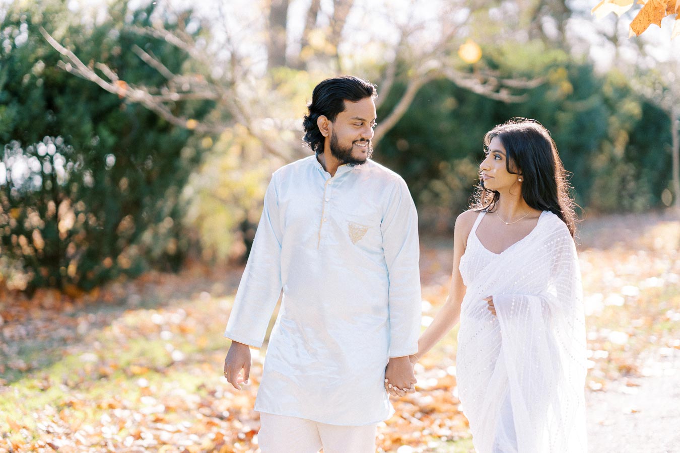 A couple walking hand in hand in a park, wearing traditional Indian attire, surrounded by autumn foliage.