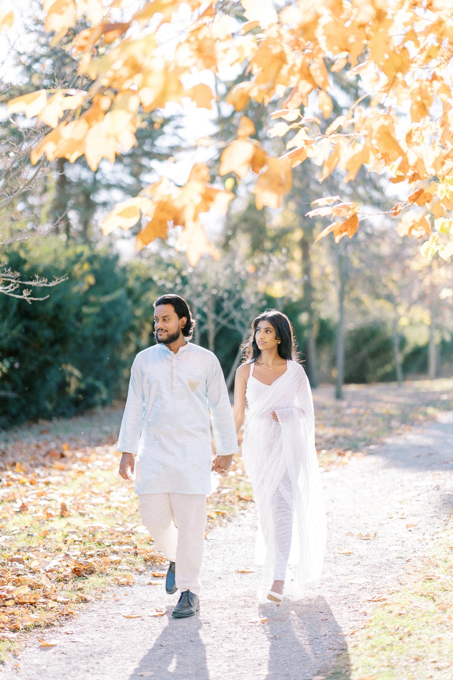 A couple holding hands and walking through a park with autumn leaves, dressed in elegant white attire.
