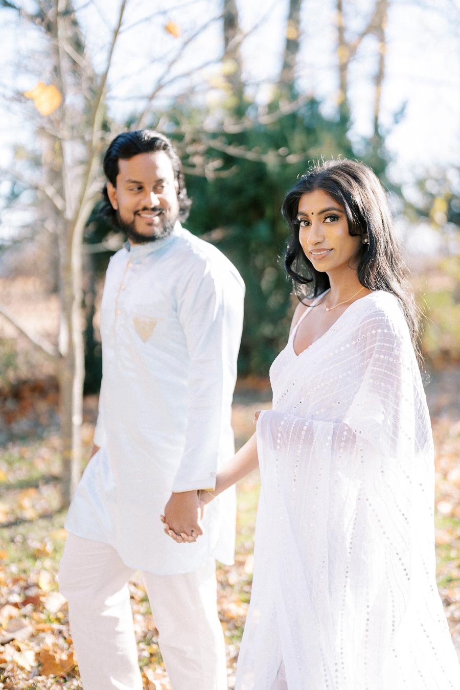 Couple in traditional attire holding hands, walking outdoors surrounded by autumn leaves and trees.