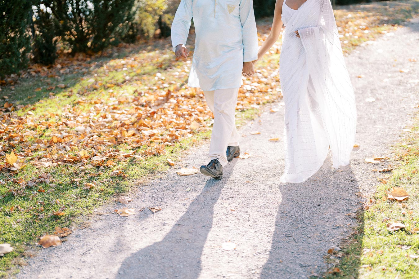 A couple dressed in traditional attire walking hand-in-hand on a sunlit path covered with autumn leaves, showcasing a romantic outdoor setting.