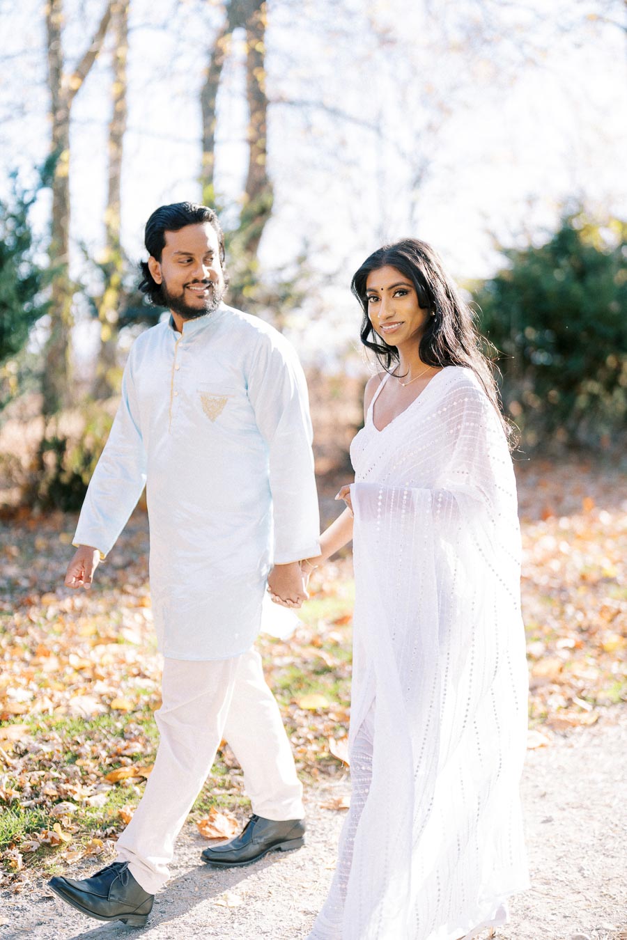 Couple in traditional attire walking hand in hand through a sunlit autumn path, surrounded by trees and fallen leaves, creating a serene and romantic atmosphere.
