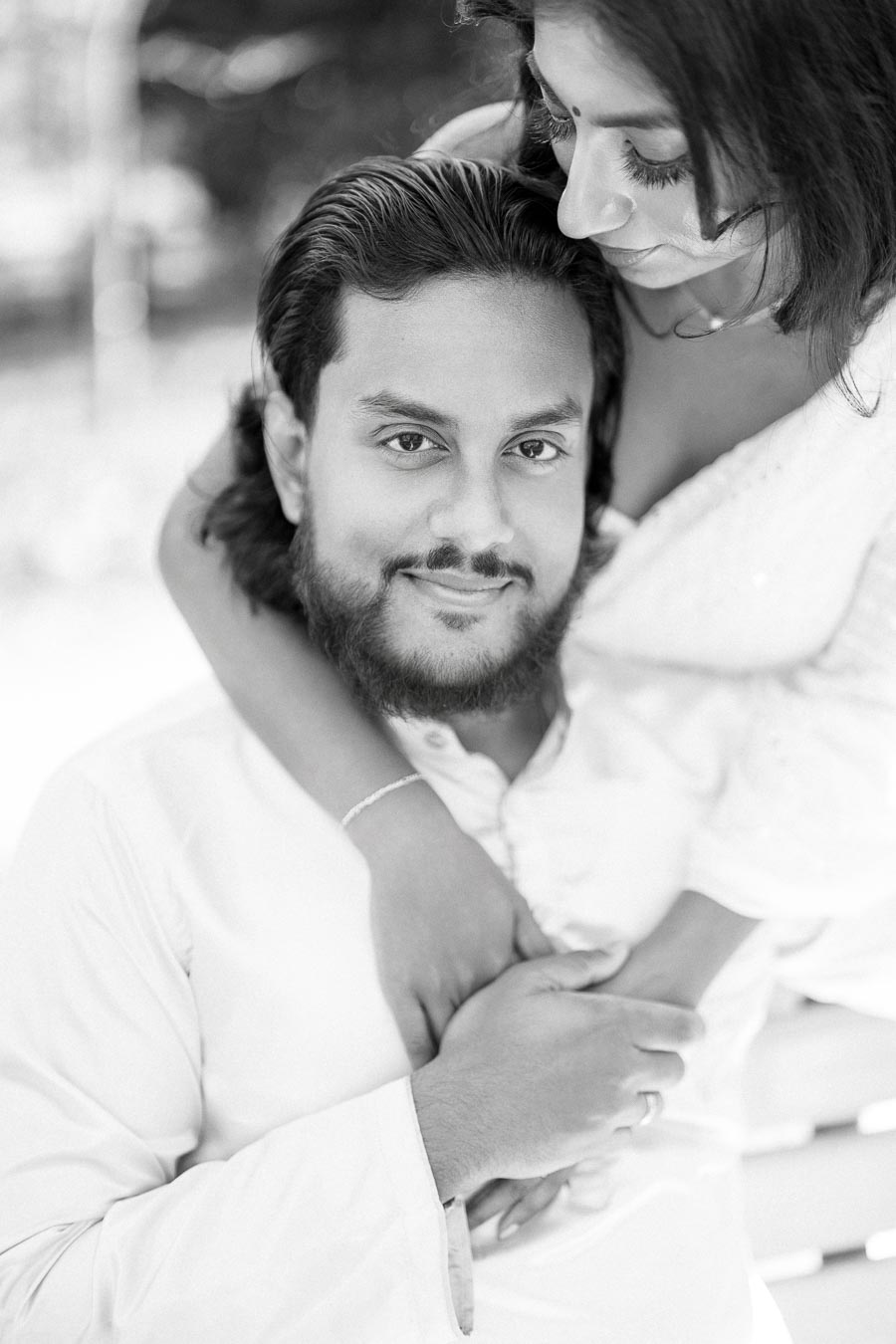 A black and white photo of a couple embracing, with the woman gently hugging the man from behind. The man is gazing at the camera with a slight smile, while the woman looks down affectionately. The photo captures a moment of tenderness and closeness.