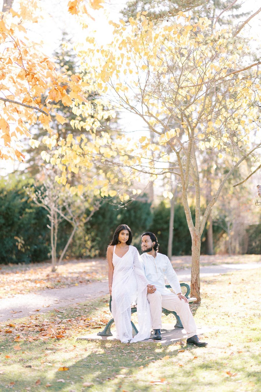 Couple in white attire enjoying a sunny autumn day on a park bench surrounded by golden foliage.