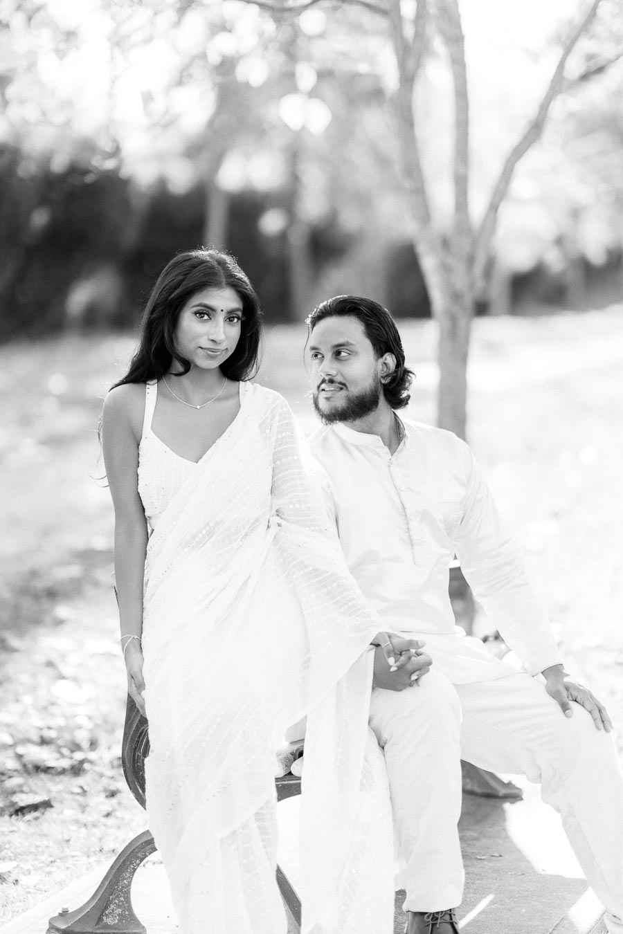Black and white photo of a couple in traditional attire, seated outdoors on a bench, with soft sunlight filtering through trees in the background.