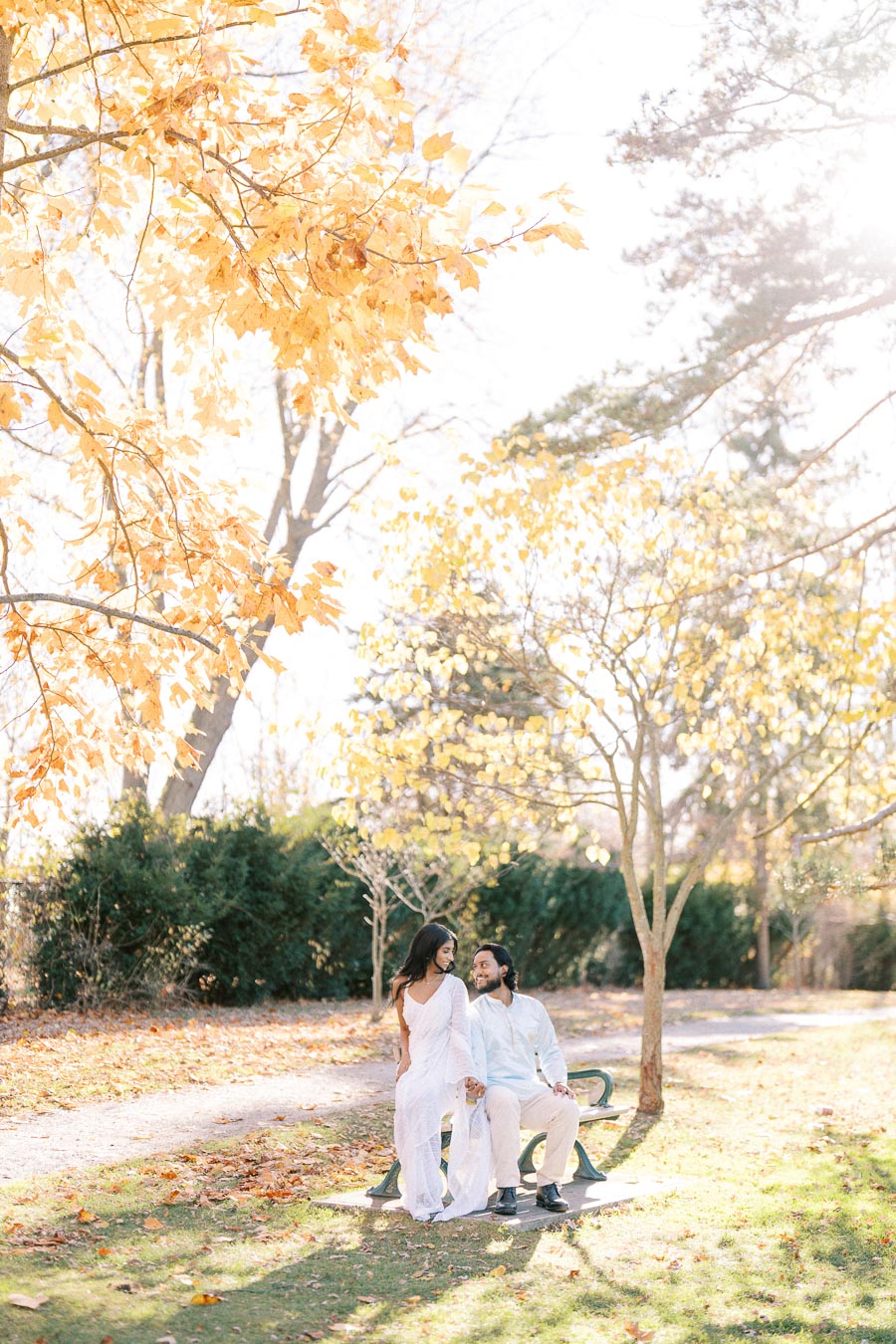 A couple sitting on a park bench surrounded by autumn leaves, with golden foliage and sunlight creating a warm, romantic atmosphere.