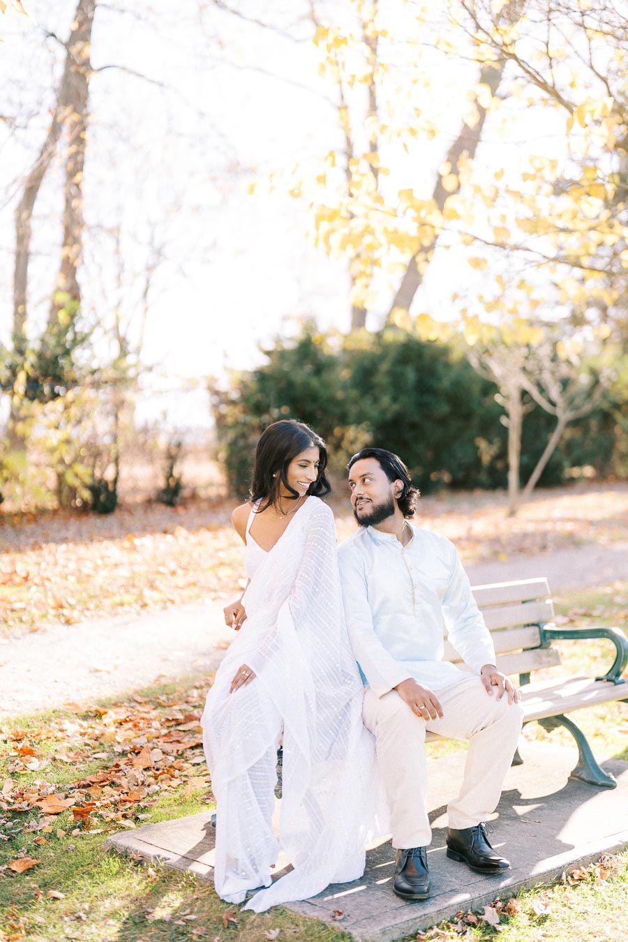 A couple enjoying a sunny autumn day on a park bench, surrounded by fallen leaves and vibrant trees, with the woman in a white saree and the man in a light blue outfit, sharing a joyful moment together.