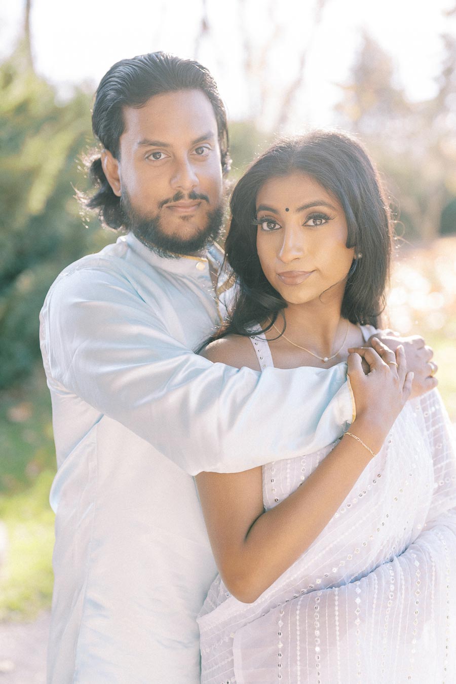 Couple embracing in a serene outdoor setting, with woman wearing a traditional white sari and man in a light-colored kurta.