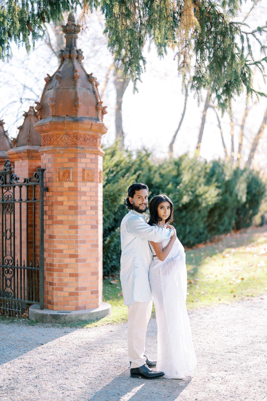 Couple embracing in a garden setting with a brick column and wrought iron gate, surrounded by greenery and soft sunlight filtering through trees.