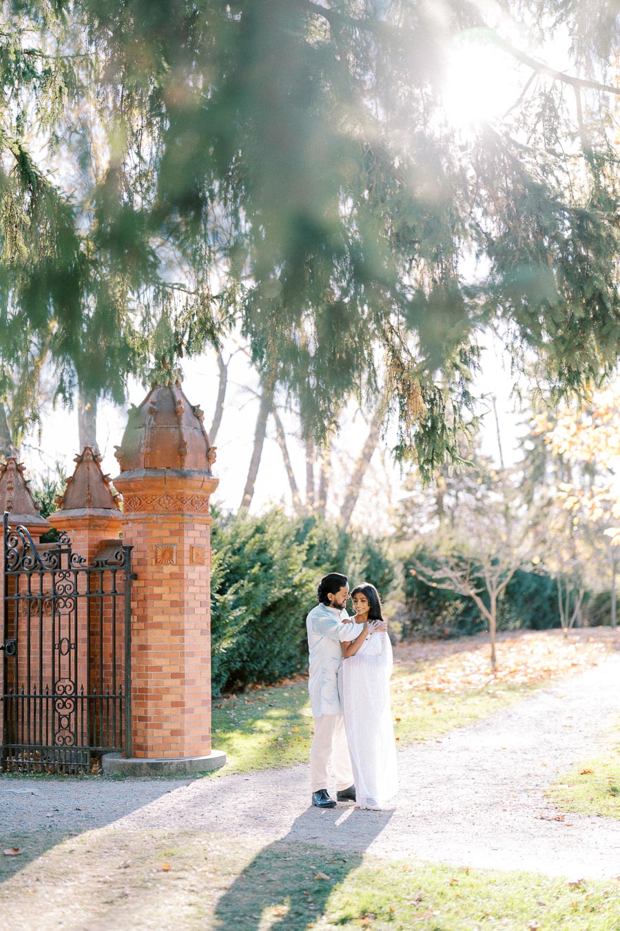 A couple embraces under dappled sunlight in a garden setting, with lush green trees and ornate brick pillars in the background.