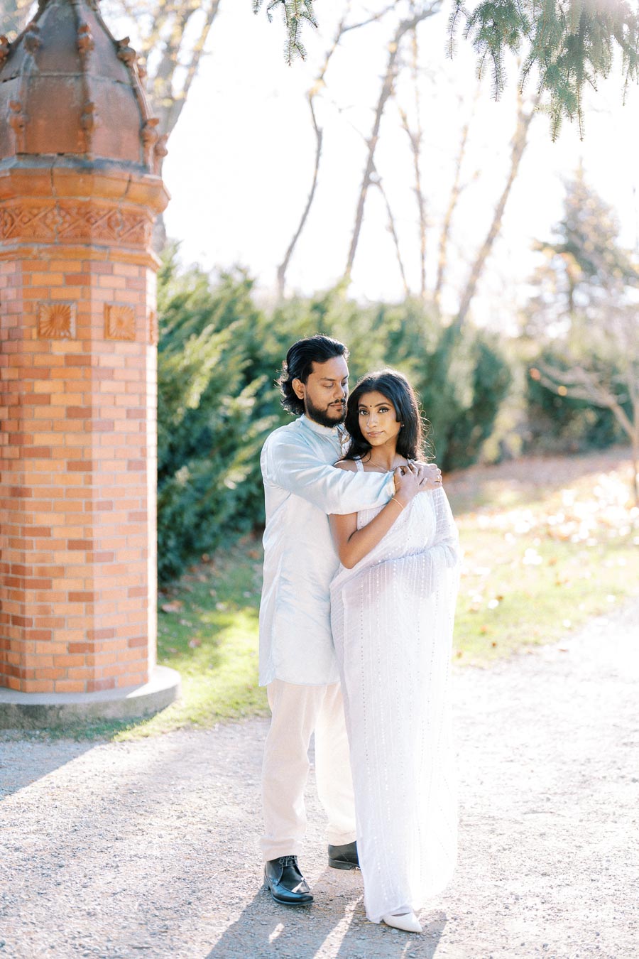 A couple embraces tenderly in a sunny garden setting, with greenery and a decorative brick pillar in the background. The man wears a traditional light-colored outfit, while the woman is draped in a flowing white saree, evoking romance and elegance.
