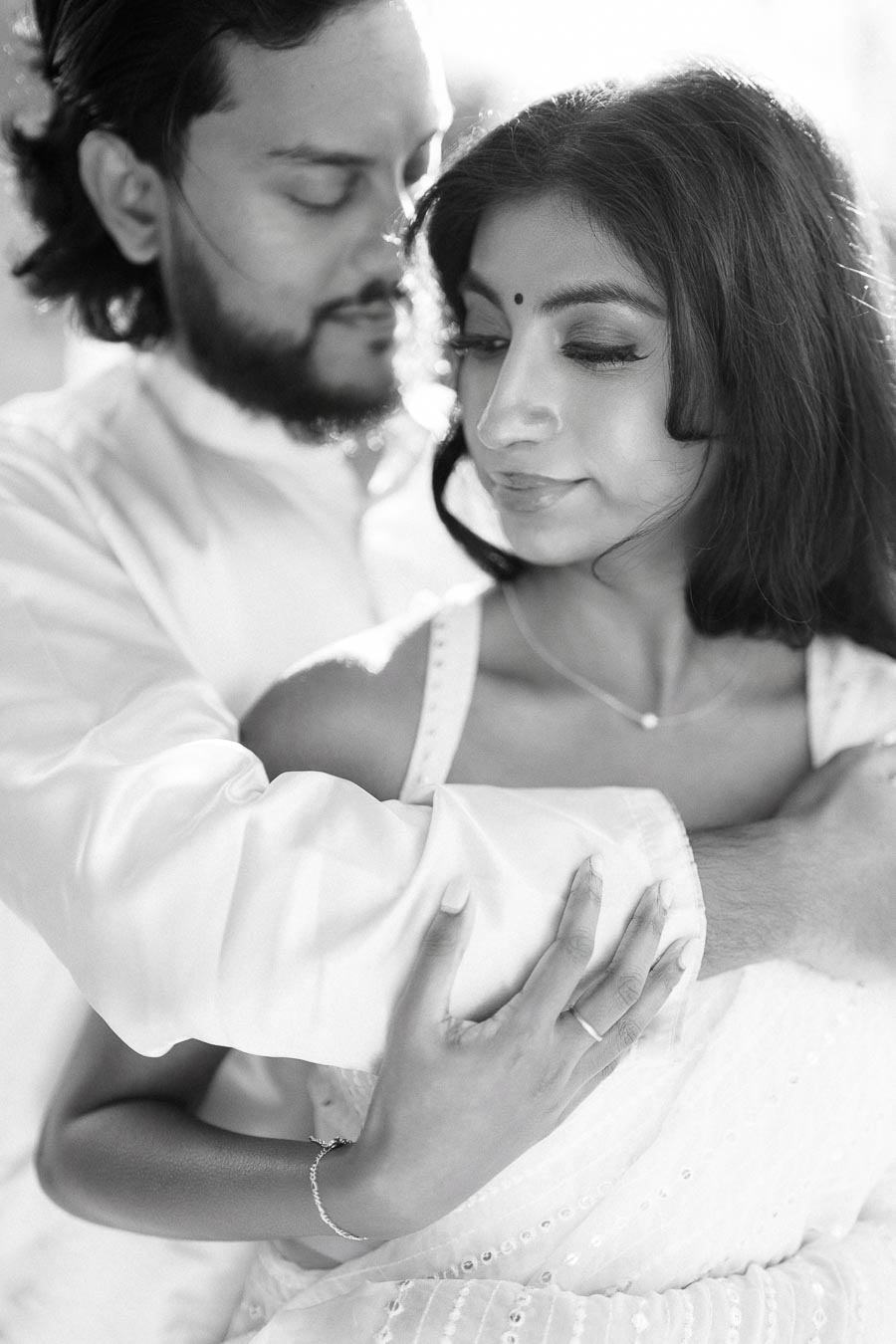 A romantic black and white photo of a couple embracing, with the woman looking downward and the man gently holding her shoulders from behind. Both appear serene and affectionate.