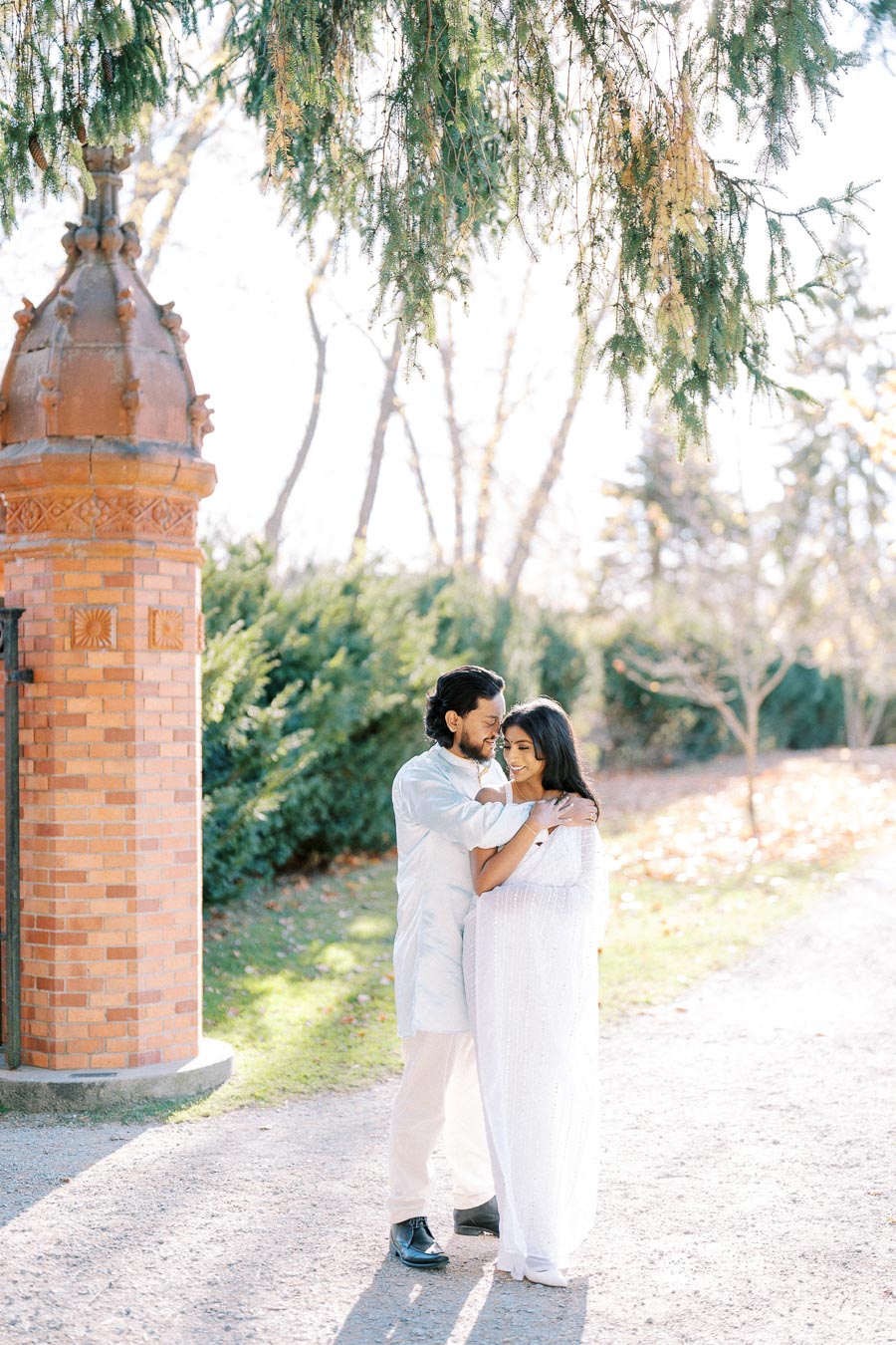 A couple embracing under a tree in a sunlit park with a brick gazebo in the background, wearing light-colored clothes.