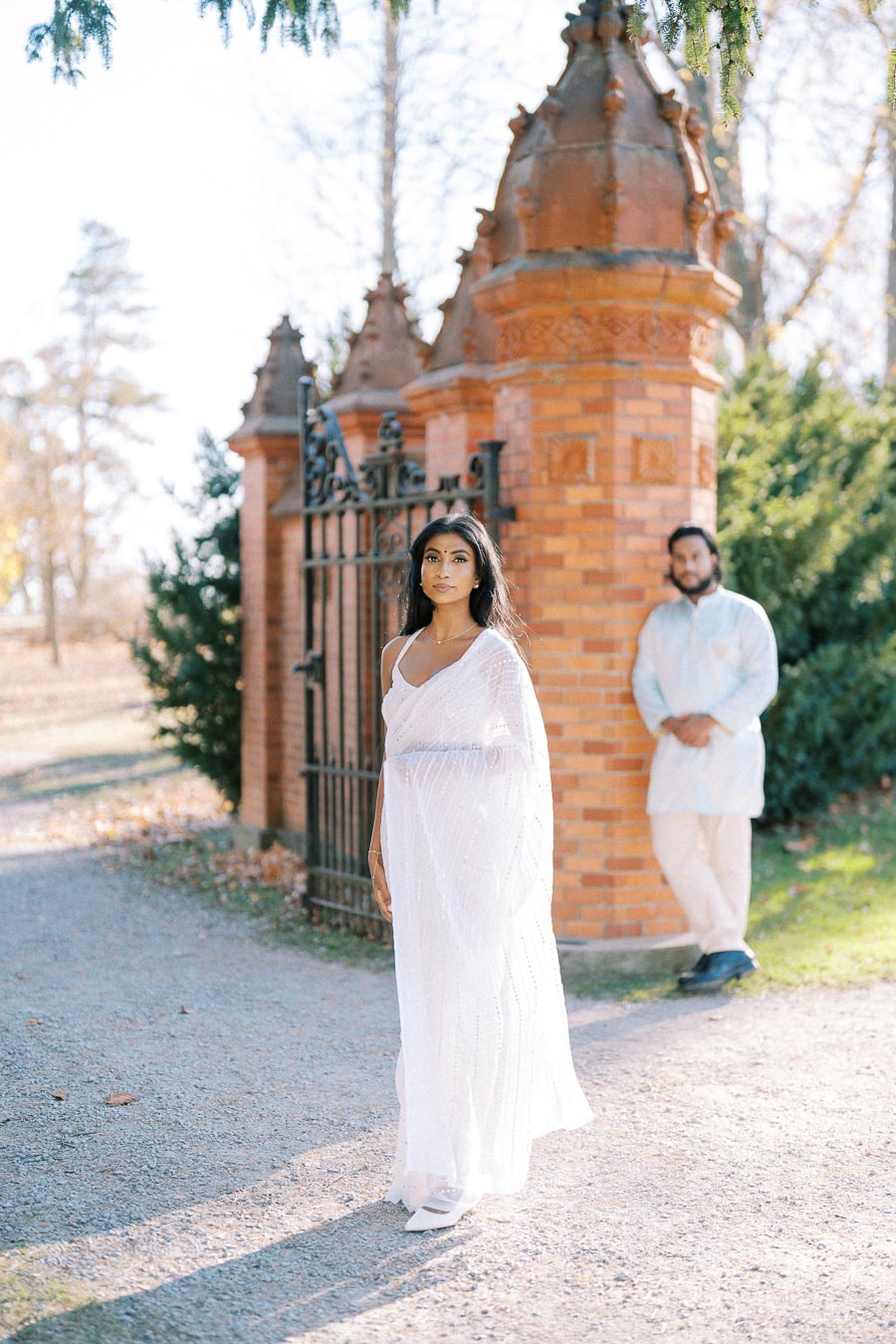 Woman in elegant white sari standing in front of an ornate iron gate and brick pillars, with a man in traditional attire in the background, outdoors on a sunny day.
