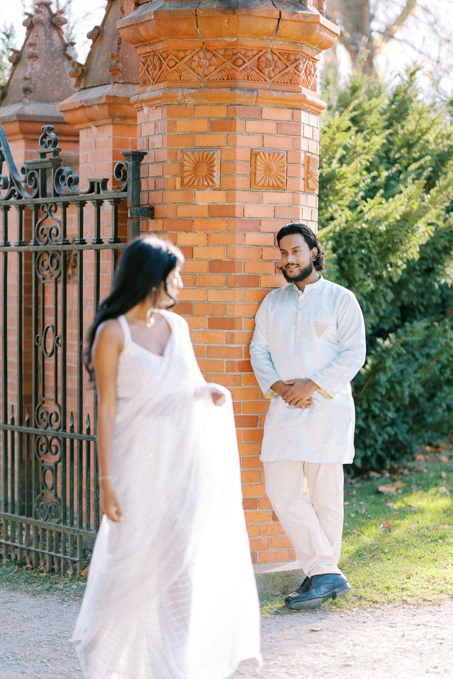 Couple in traditional attire by a brick wall and wrought iron gate, basking in sunlight with lush greenery in the background.