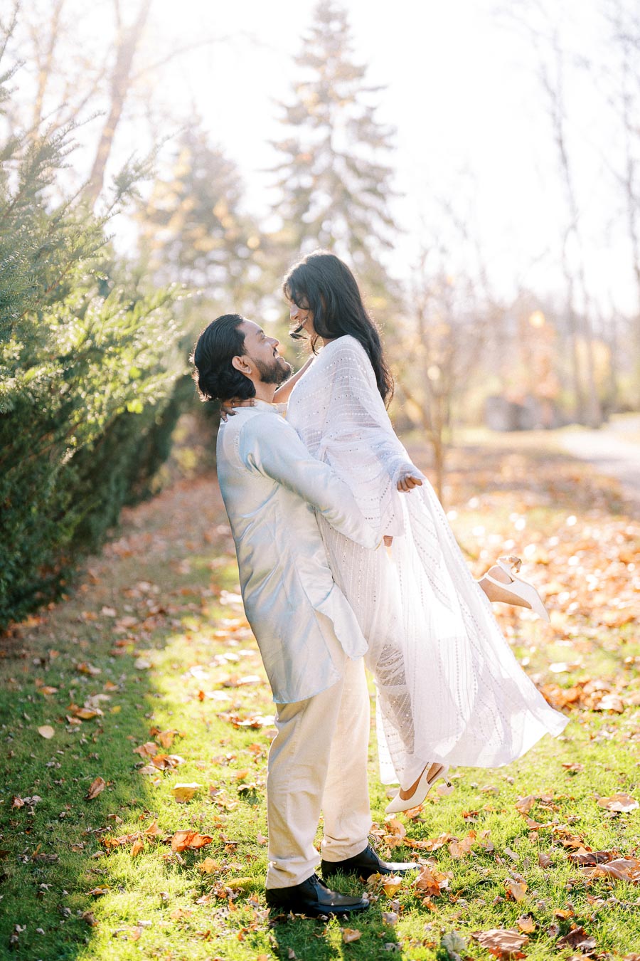 A couple in elegant white attire enjoying a romantic moment outdoors on a sunny autumn day, with fallen leaves on the grass and a blurred background of trees.