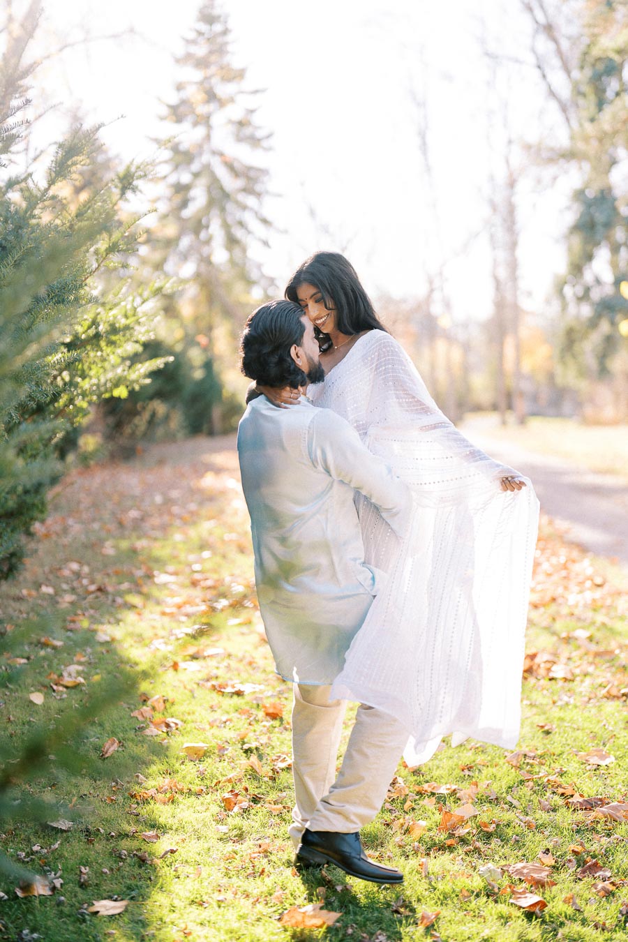 A couple enjoying a joyful moment outdoors, with the man lifting the woman as they both smile brightly, surrounded by autumn foliage and sunlight filtering through trees.