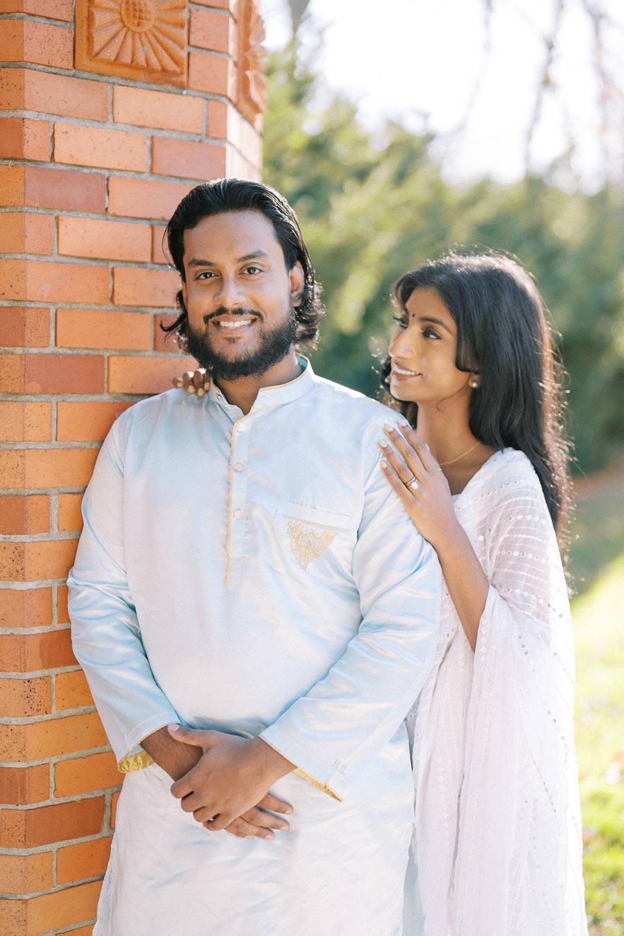 Happy couple in traditional attire standing by a brick pillar, with the woman embracing the man from behind, outdoors in daylight.