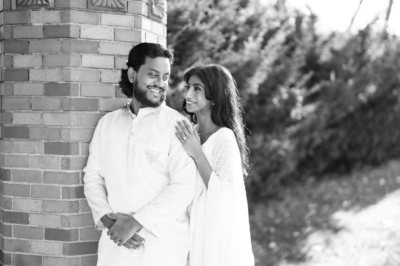 A happy couple wearing traditional attire smiles warmly while leaning against a brick column, surrounded by lush greenery, capturing a romantic and serene moment.