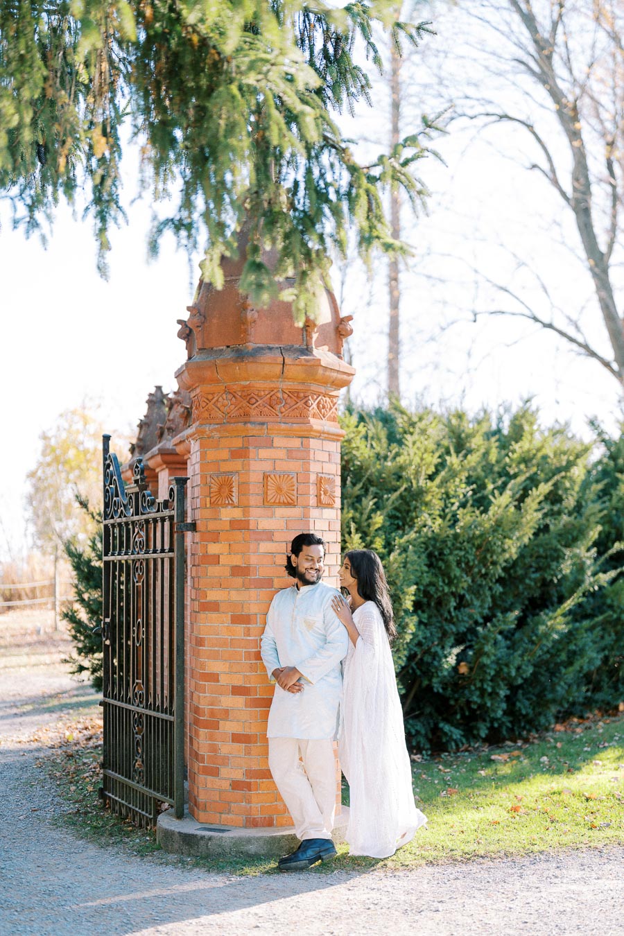 A couple in traditional attire poses affectionately by a brick pillar and ornate gate, surrounded by lush greenery and sunlight filtering through the trees.