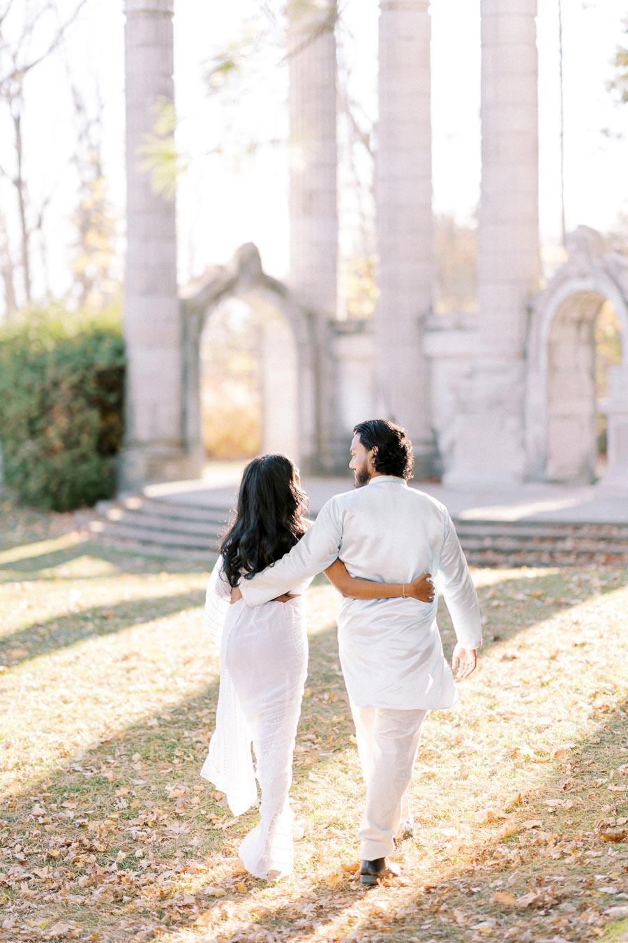 A couple walking hand in hand in a park with ancient stone columns and autumn leaves on the ground, both dressed in white.