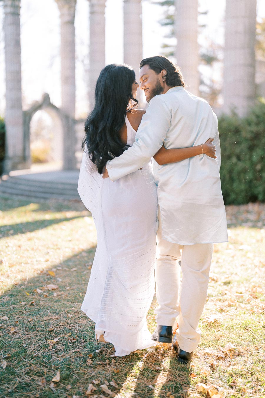 A couple dressed in white traditional attire walking arm in arm through a sunlit park, surrounded by tall columns and fallen leaves, creating a romantic and serene scene.