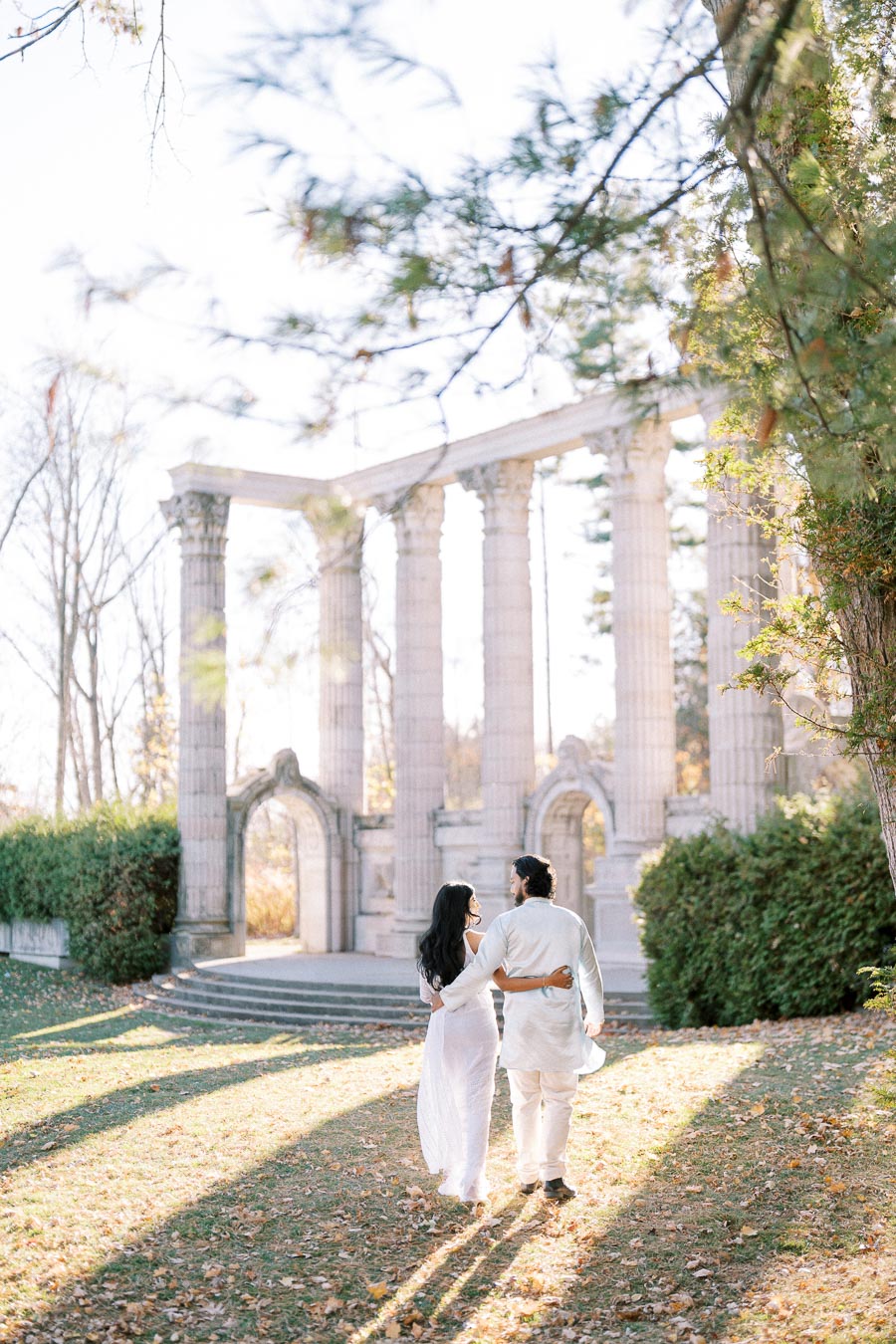 Couple in white outfits walking through autumn leaves towards historic stone columns in a park setting.