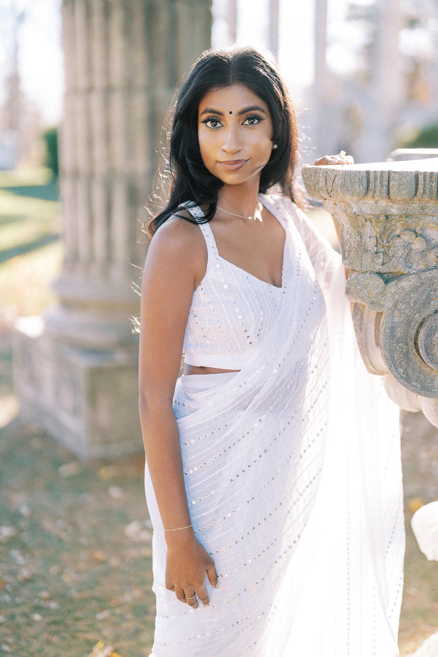 Young woman in a light-colored traditional sari standing by a stone pillar, captured in an outdoor setting with soft natural lighting.