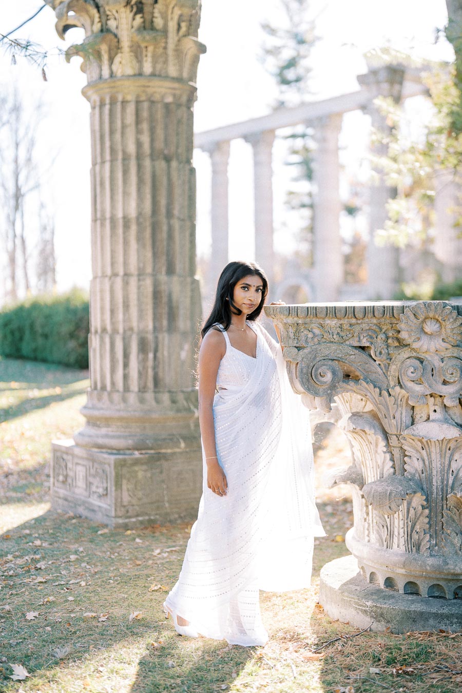 A woman in a white sari standing by an ancient stone column, surrounded by a serene outdoor setting with sunlight filtering through.