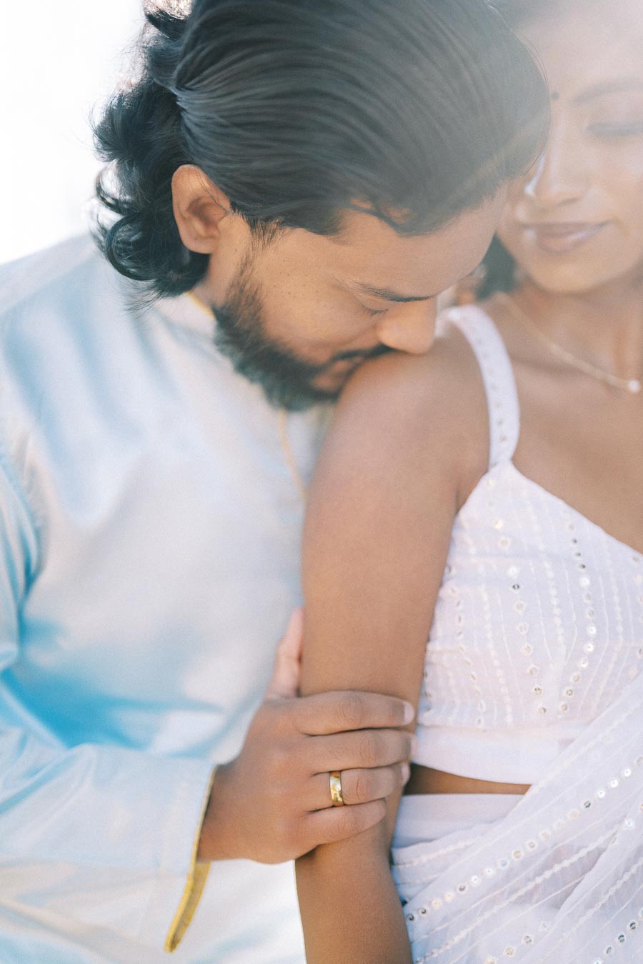 Couple embracing in soft romantic moment, dressed in elegant traditional attire, highlighting a gold ring on the man's finger.