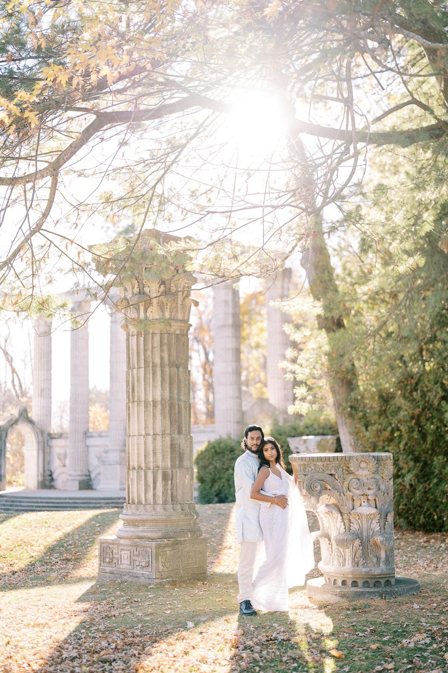 A couple embracing in a sunlit garden, surrounded by ancient stone columns and lush greenery, with autumn leaves scattered on the ground.