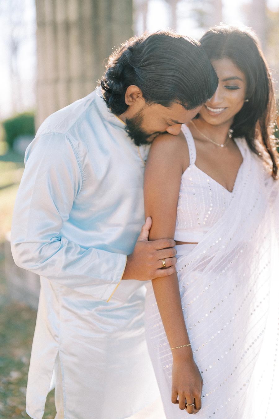 A couple in elegant traditional attire sharing an intimate moment outdoors, with the man gently embracing the woman’s arm and displaying feelings of affection, bathed in soft natural light.