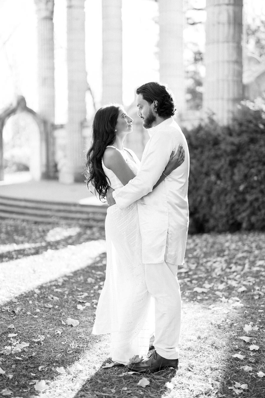 A couple in white attire embracing outdoors in a park with ancient pillars in the background, showcasing a romantic and timeless moment.