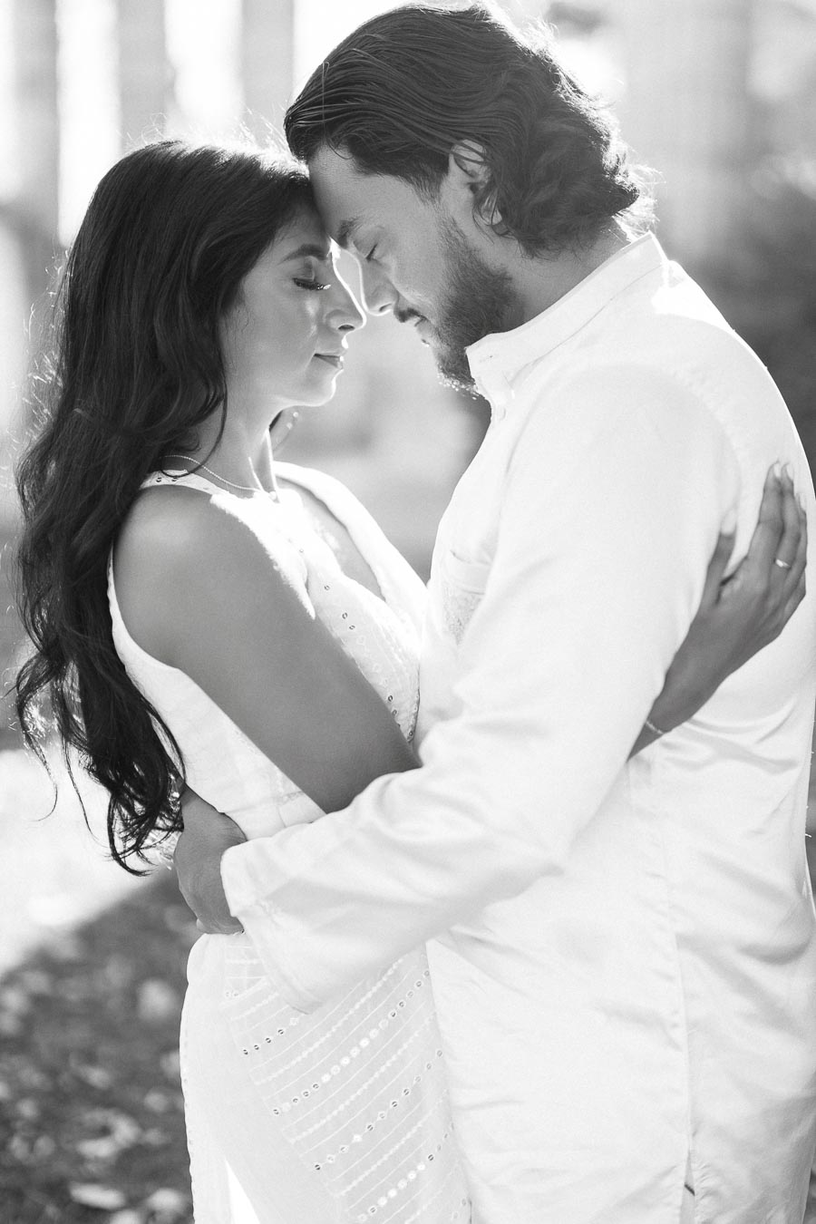 A black and white photo of a couple embracing, wearing white outfits, with their foreheads touching in a serene outdoor setting.