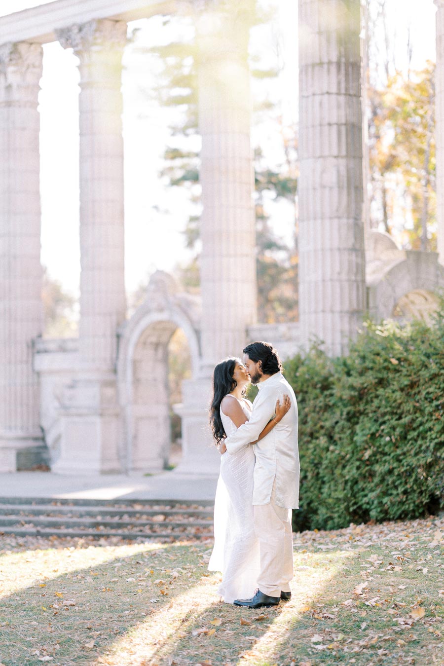 A couple embracing in an outdoor setting with historic stone columns in the background, surrounded by greenery and dappled sunlight.